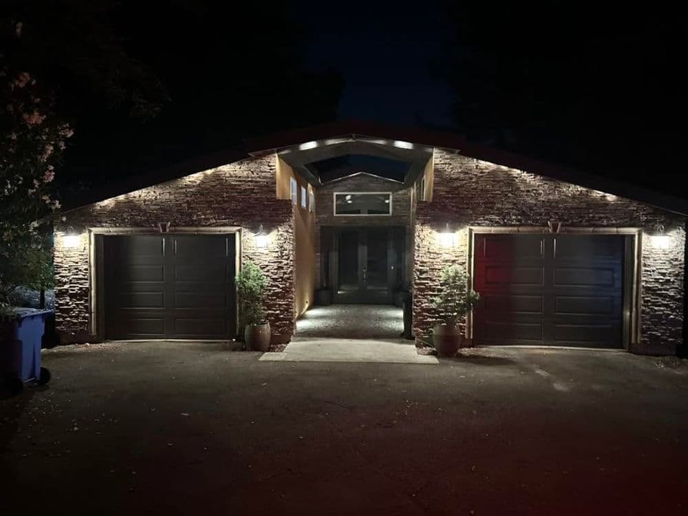 Modern stone house exterior at night with illuminated driveway and garage doors.